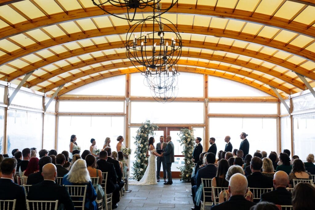 Ceremony indoors with open floral arbor of all white flowers and wedding party standing at the front with the bride & groom